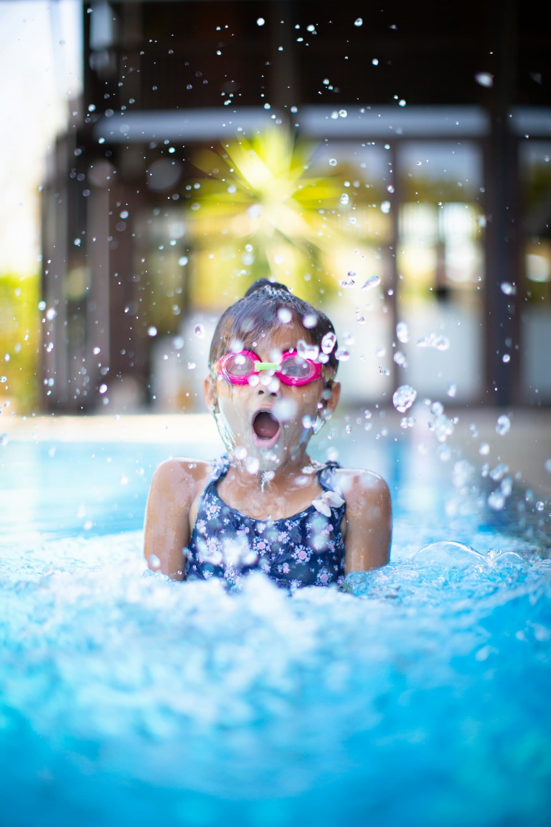 Menina de touca e óculos de natação sorrindo na piscina, com água espirrando